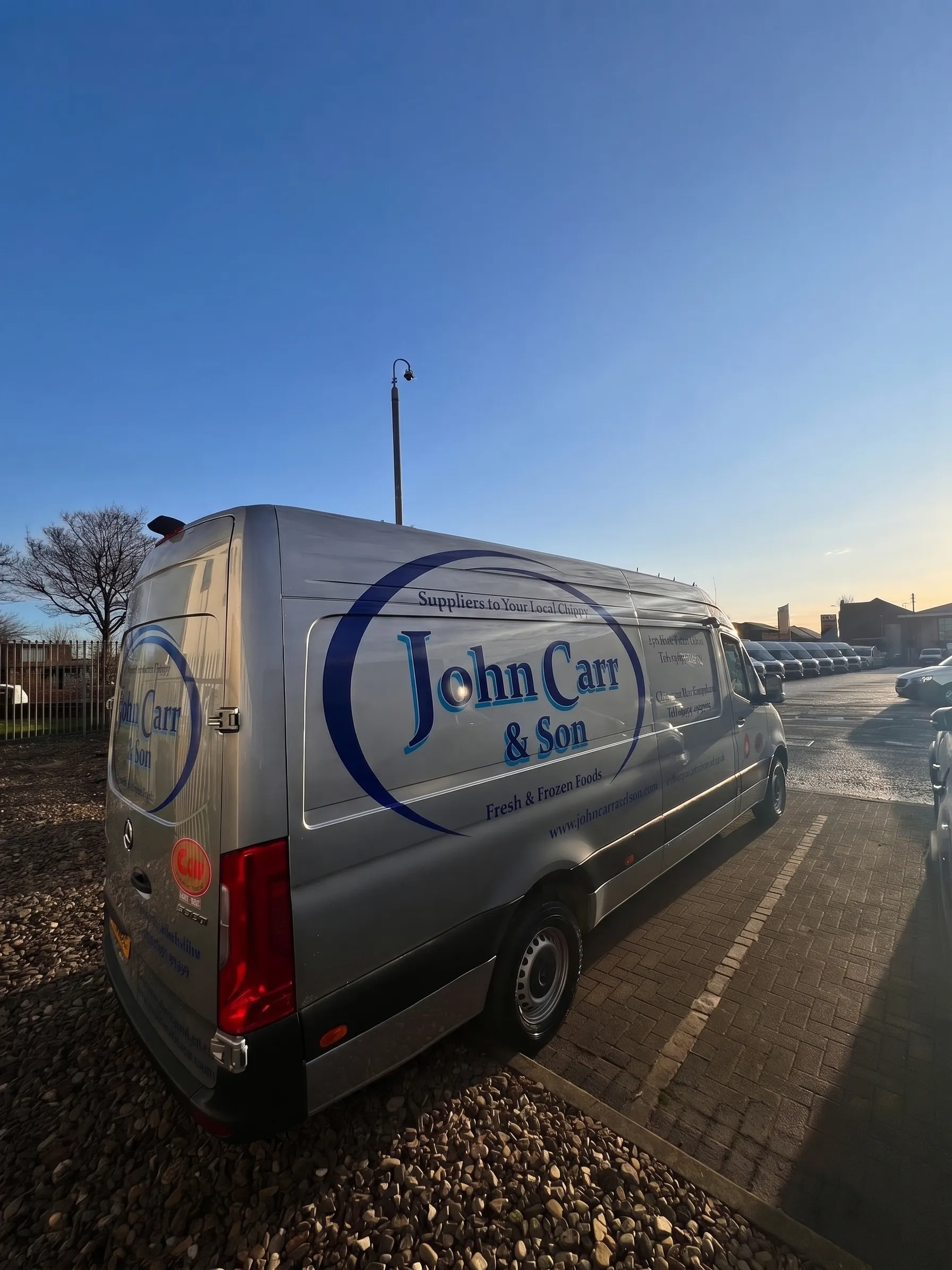 A silver John Carr & Son delivery van parked on block paving under a wide, cloud-streaked sky with low golden light on the horizon. Its livery reads “Suppliers to Your Local Chippy” and “Fresh & Frozen Foods”.